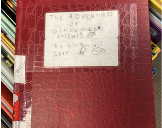 Photograph of the cover of Dillon's addition to his local Idaho library. In a child's handwriting, the cover says 'The Adventures of Dillon Helbig’s Crismis by Dillon his self'