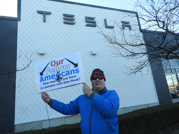 Protester in front of a Tesla building holds a sign reading, “Our Fellow Americans: How much do you have in common with Elon Musk?” The person is dressed warmly in a blue jacket, sunglasses, and a beanie. The Tesla logo is visible on the building behind them, with bare tree branches framing the side.