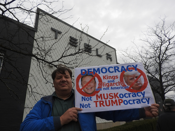 Protester in front of a Tesla building holds a sign reading, “DEMOCRACY: Kings and Oligarchs are not American. Not MUSKocracy, Not TRUMPocracy,” with images of Elon Musk and Donald Trump crossed out. The person wears a blue jacket, standing against a gray, overcast sky, with bare tree branches framing the scene.