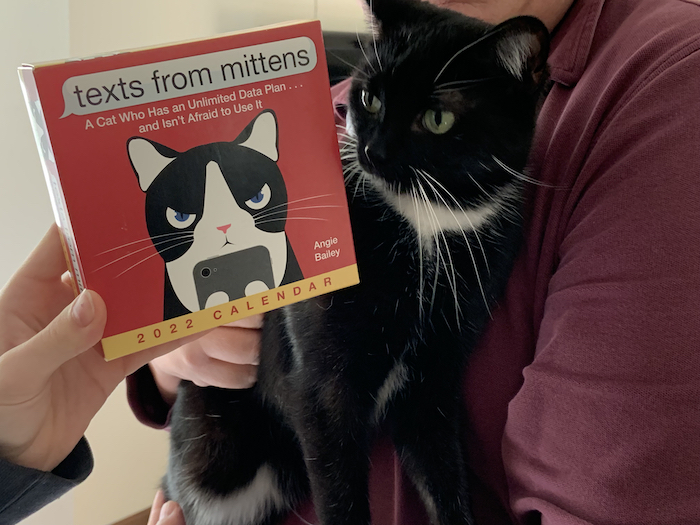Photograph of a black cat in the arms of its owner. Held next to the cat is a page-a-day calendar with the title 'texts from mittens'. The picture of the cat on the calendar box resembles the cat being held.